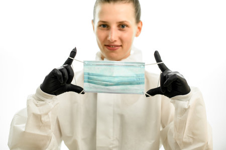 Young woman in white protective suit and black gloves hold medical mask in her hands