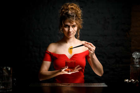 Beautiful woman professional bartender holds glass with cocktail and decorates it with orange slice.の写真素材