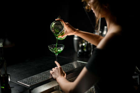 professional female bartender in dark bar pours cocktail from cup to glassの写真素材