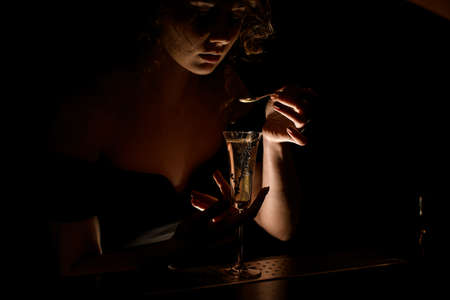 attractive girl at dark bar holds teaspoon over glass with cocktailの写真素材
