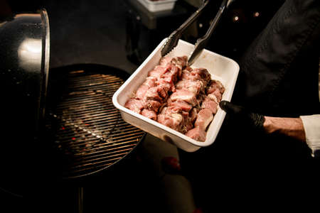 male chef in black gloves with special tongs begin to spread raw meat pieces on barbecue grillの写真素材