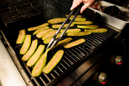 Close-up of zucchini slices on grill grid and fried on fire. Cook's hand holds tongs and flips slicesの写真素材