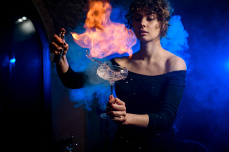 beautiful female bartender holds glass with splashing drink, sprinkles and make fire over it.
