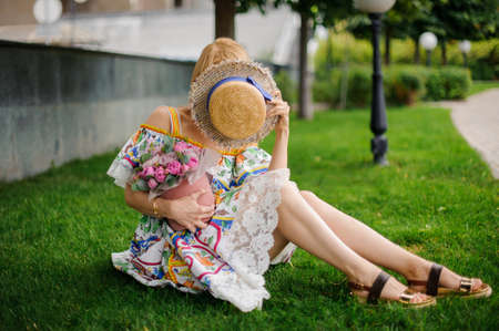 young woman sits on lawn with bouquet and covers her face with hatの写真素材