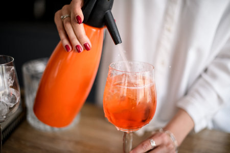 Close-up of glass with orange drink which woman is adding liquid using siphon.