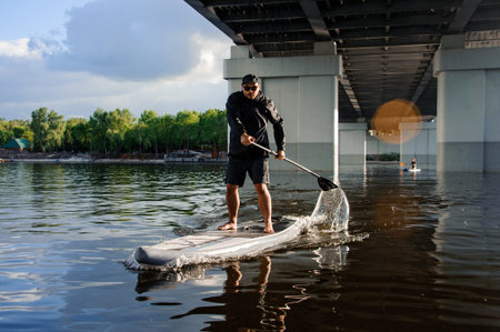 View on man which stands on sup board and swim on the river under bridge.