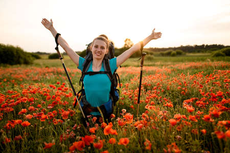 young smiling woman with backpack stands on poppy field with hands upの写真素材