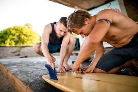 two young men carefully prepares surfboard for swimming