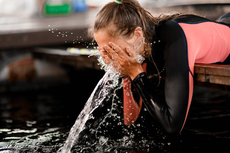 young woman wearing wetsuit washes her face with water and splashes and water droplets around her