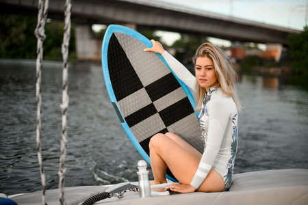 young attractive woman in swimsuit sits on boat and holds surfboard with her handの写真素材