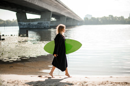 young blonde woman walks along the river bank with wakeboard in her hand