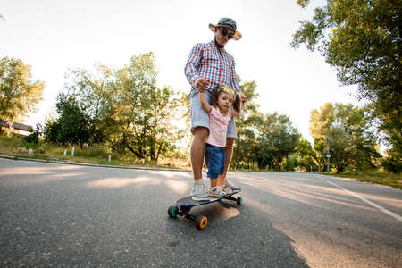 father with daughter stands on skateboard on the road in the parkの写真素材
