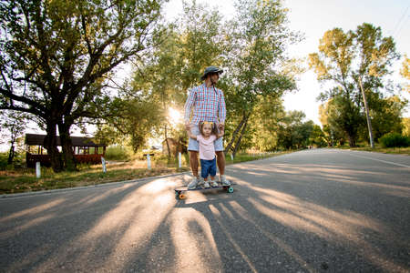 Adult man is riding skateboard with his little daughter on the road in the parkの写真素材
