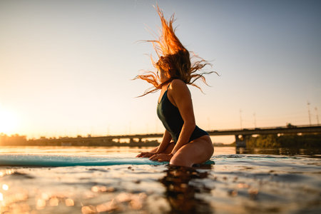 side view of attractive woman with long flowing hair sitting on surfboard against backdrop of rising...