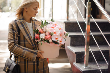 beautiful young blonde woman in coat holds flower arrangement of roses and orchids.