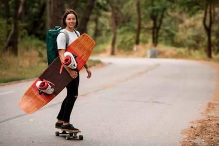 side view of girl riding skateboard with wakeboard in her hand and backpack on her shoulders.の写真素材