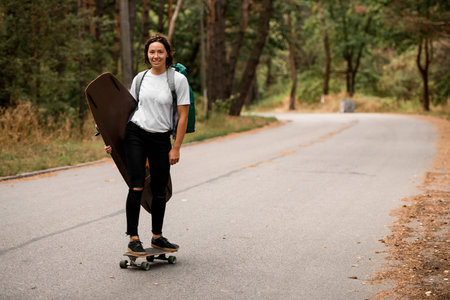 sportive young woman riding skateboard with wakeboard in her hand and backpack on her shoulders.
