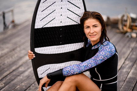 beautiful woman in black swimsuit sitting on wooden pier and holding surfboard with her hand