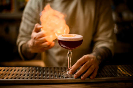 male bartender sets to fire on wine glass with foam cocktail on bar counterの写真素材