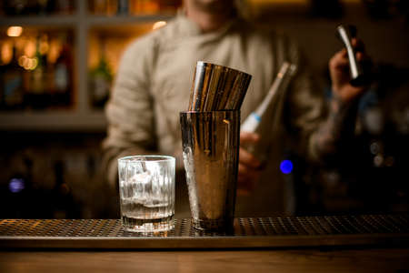 old fashioned glass with ice cubes and shaker stand on bar with bartender in backgroundの写真素材