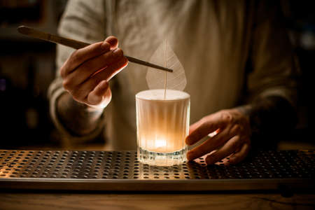Man holds tweezers with decorative leaf and carefully decorate glass of alcoholic cocktailの写真素材