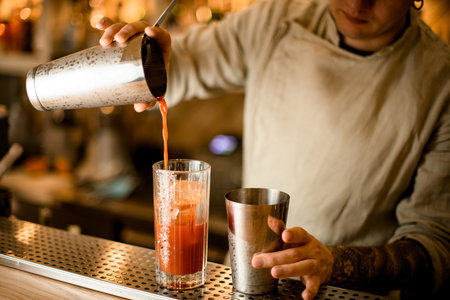 man bartender holds steel shaker glass and pours cocktail from it into other glass.