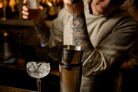 Man bartender gently pours drink from metal jigger into shaker on bar counterの写真素材