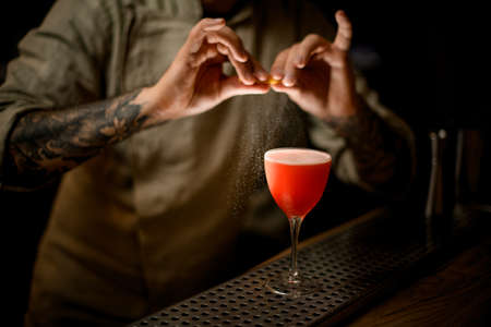 barman gently holds lemon peel and sprinkles on glass with bright cocktail.の写真素材
