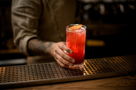 mans hand holds glass with cold drink with ice cubes, decorate with orange slice