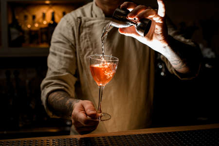 male bartender holds with hand wine glass with drink and adds liquid from jiggerの写真素材