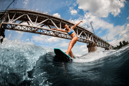 woman standing on the wake board on high wave on the background of the city bridge.