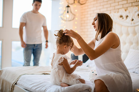 beautiful young adult woman straightens the hair of little cute girl sitting on the bedの写真素材