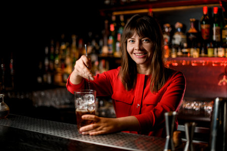 smiling woman bartender stands behind bar and mixes cocktail in glass with spoonの写真素材