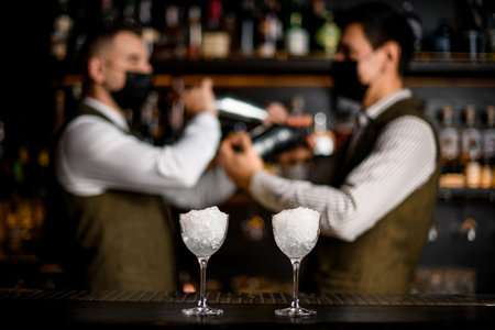 close-up of two glasses on bar counter and bartenders with cocktail shakers in backgroundの写真素材