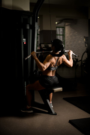 Back view of athletic woman pulls on simulator. Concept of sports, gyms, sportswear, fitness.