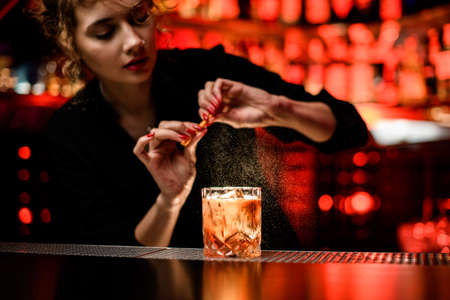 woman bartender gently sprinkles on glass with cold alcoholic cocktail on the bar counterの写真素材