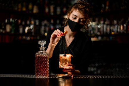 view on woman bartender in mask who holds glass of cocktail and sprinkles on itの写真素材