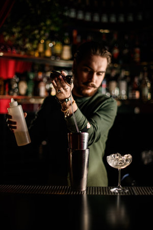 male bartender skilfully pours liquor from jigger into steel shaker cup on bar counter