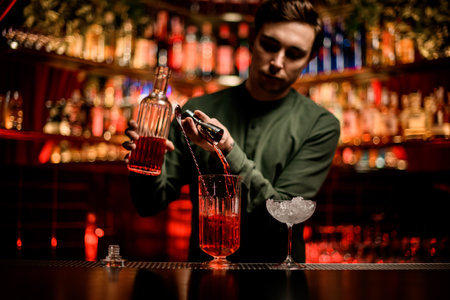 young man bartender pouring bright alcoholic beverage into mixing cup on bar counterの写真素材