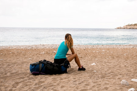 rear view of young woman tourist sitting on backpack on seashoreの写真素材