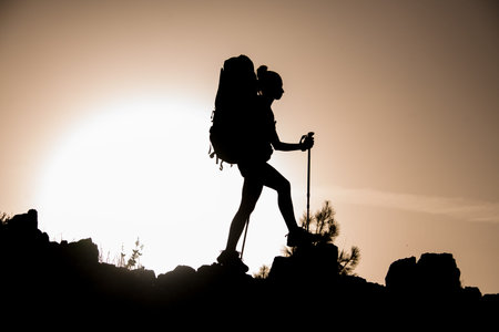 silhouette of woman with trekking sticks and backpack who walks along rocky path against sky