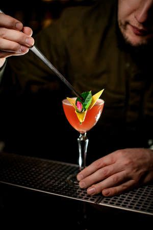 male bartender expertly decorates cocktail glass by small green leaf using tweezersの写真素材
