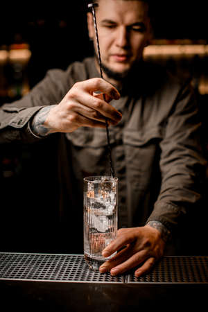 man bartender holds bar spoon and gently mixes drink in transparent glassの写真素材
