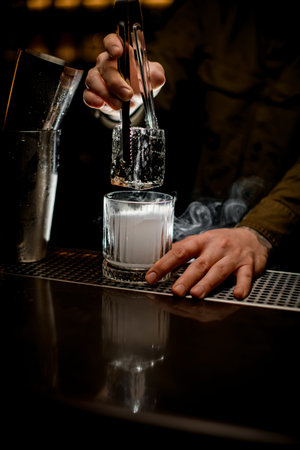 bartender neatly holds large piece of ice with tongs over steaming old-fashioned glass