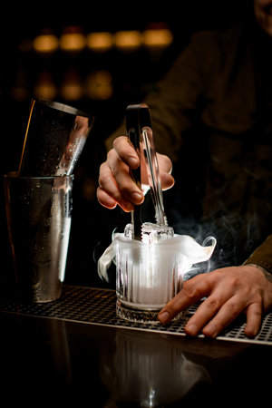 magnificent view of illuminated steaming glass in which bartender put piece of ice using tongs