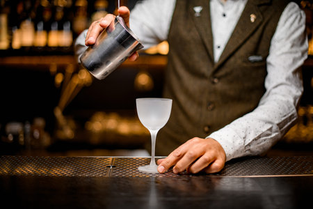 view of glass on bar over which the bartenders hand holds steel mixing cup