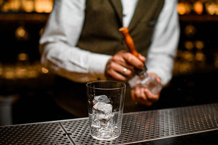 glass with ice cubes on bar. Bartender is cracking ice in the backgroundの写真素材