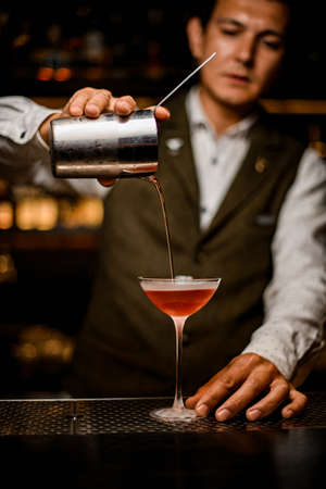 view of man bartender neatly pouring drink from mixing cup with strainer into glassの写真素材
