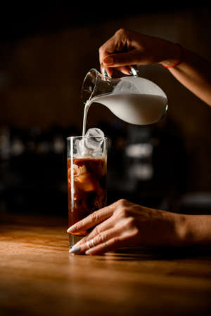 womans hand holds jug and carefully pours milk into glass with coffee and iceの写真素材