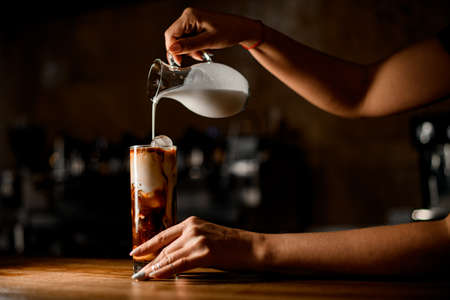 woman holds jug and carefully pours milk into glass with coffee and iceの写真素材